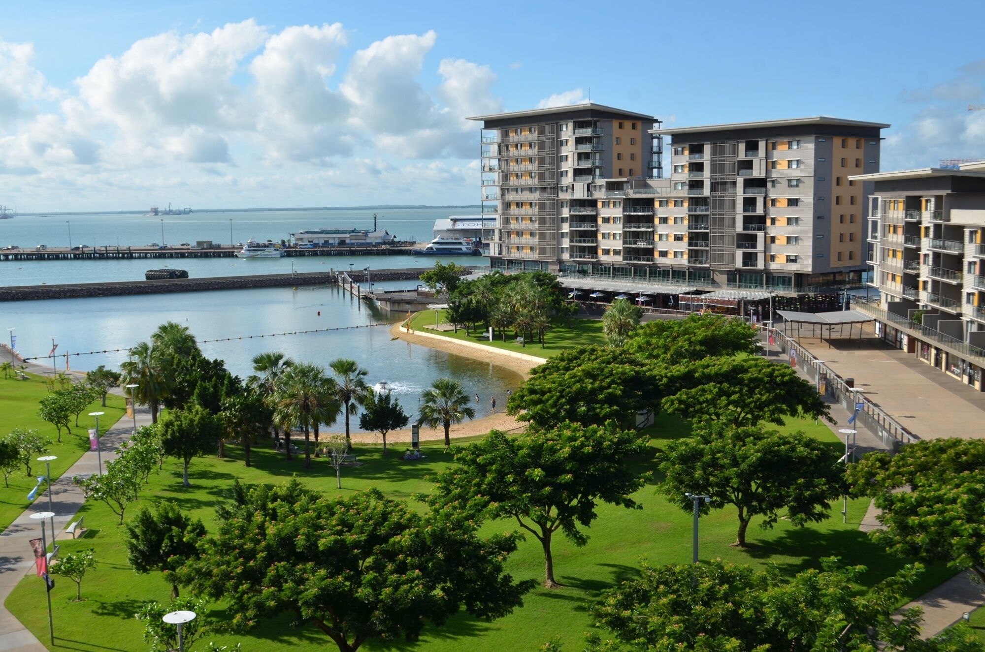 Uma área residencial costeira com edifícios de apartamentos modernos, um porto de iates e um parque bem cuidado com árvores e um pequeno lago.