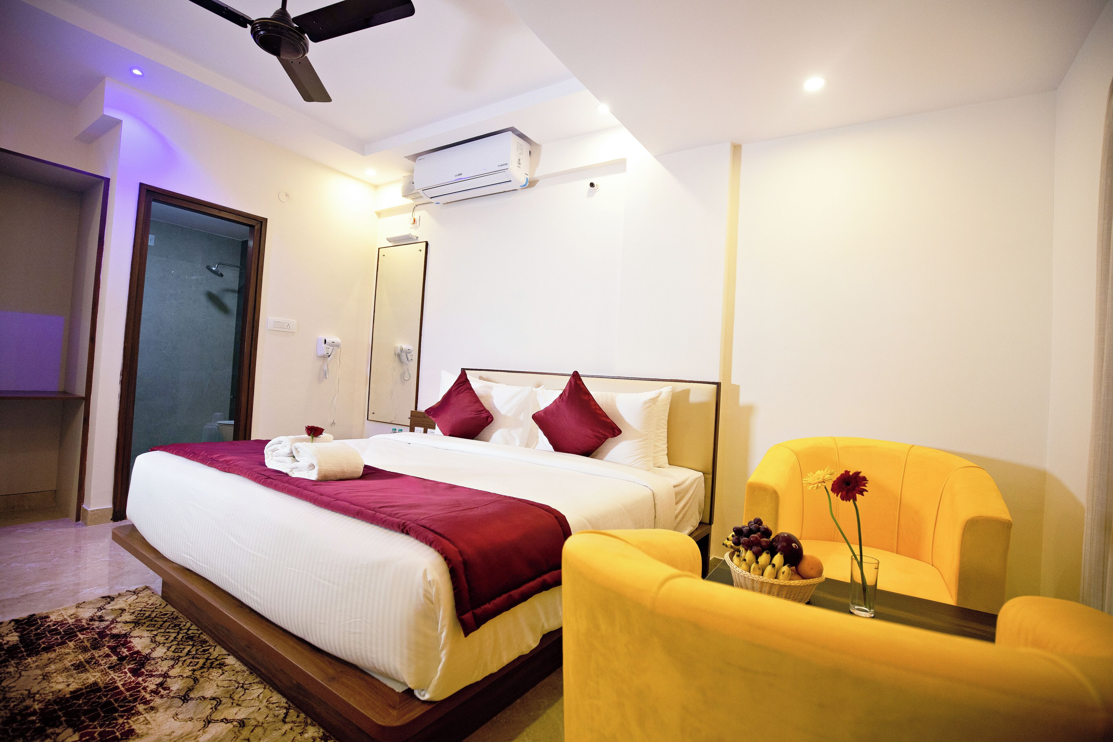 A hotel room with a bed, a chair, a small table with flowers, and an air conditioning unit.