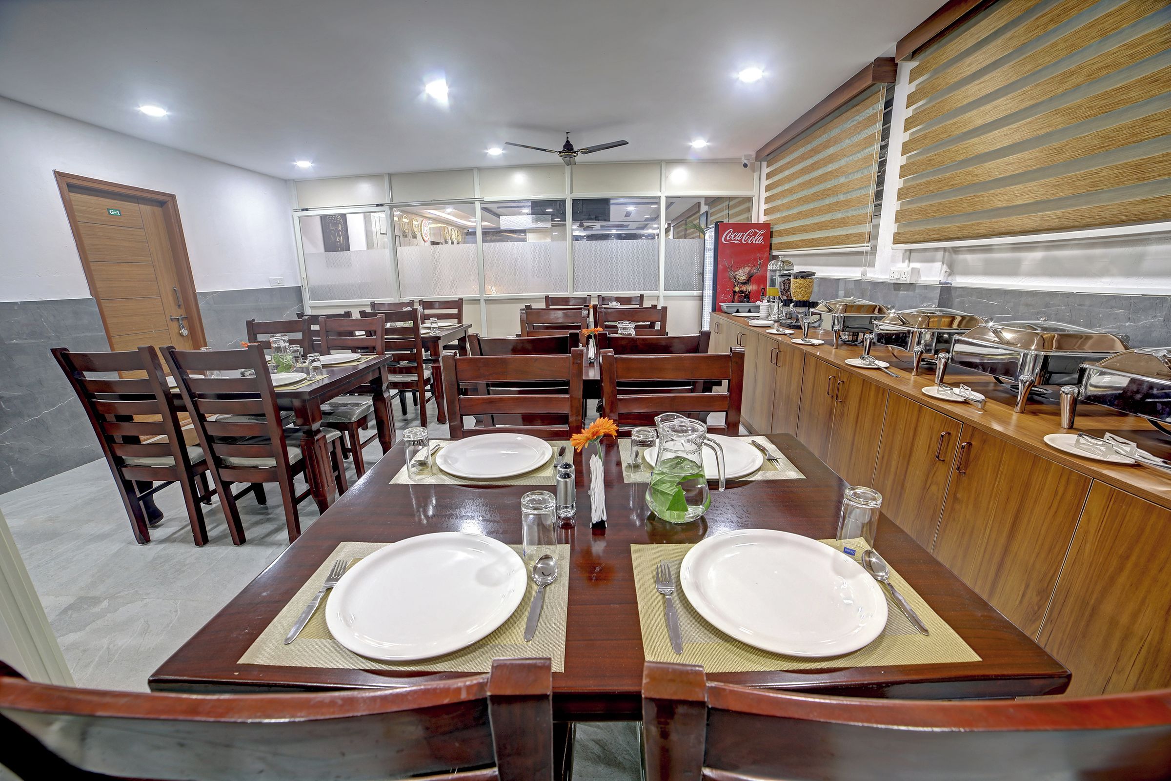 Un comedor con una mesa larga dispuesta para dos personas, rodeada de sillas, con una encimera de cocina y un refrigerador Coca-Cola al fondo.