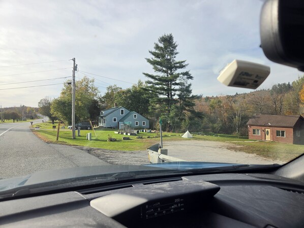 Tablero de vehículo con vista a un camino rural, una casa azul y un pequeño edificio de ladrillo.