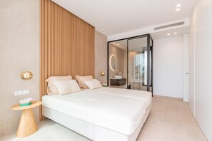 Quarto moderno com cama grande, painel de parede de madeira, box de vidro e pequena mesa redonda.
