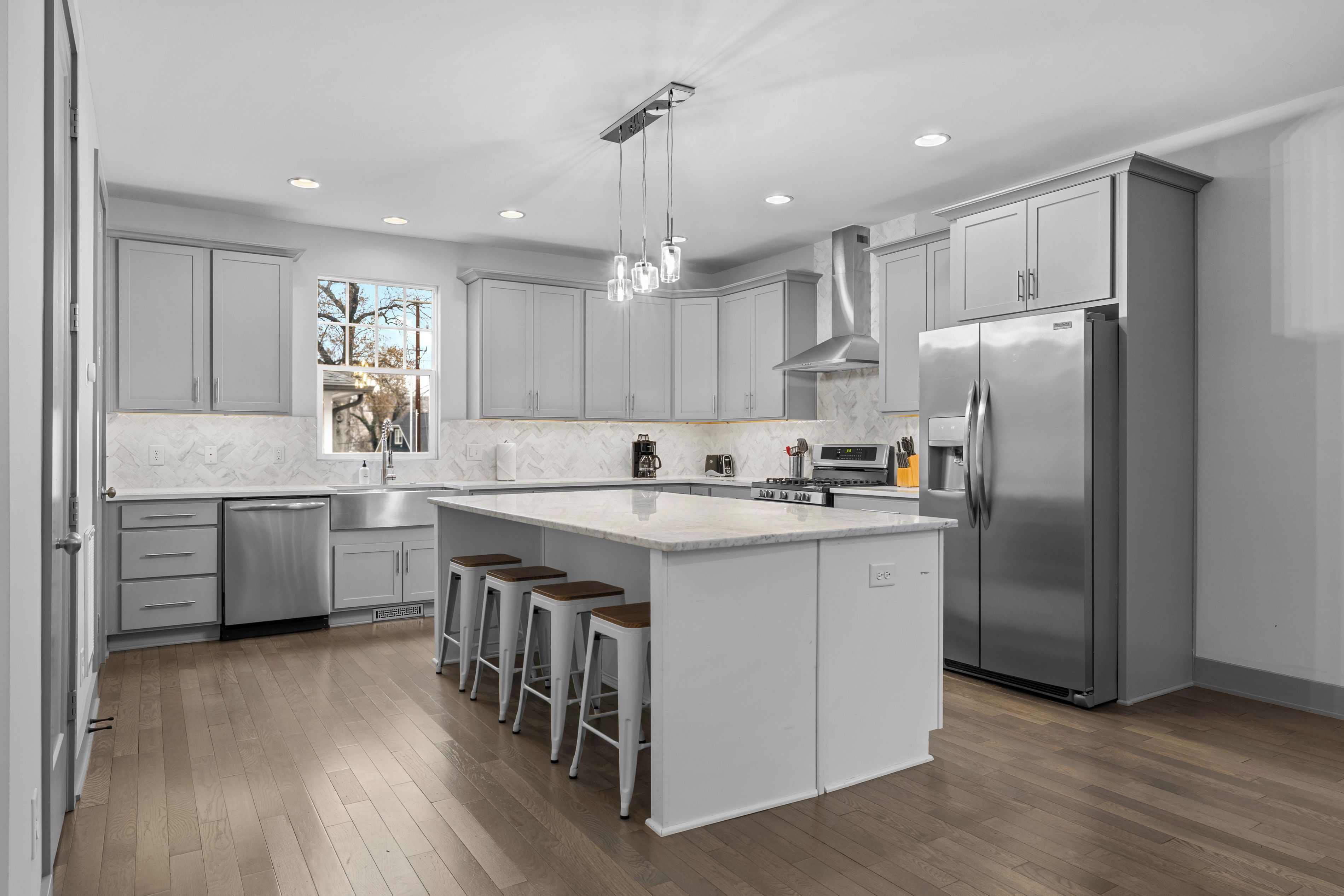 A modern kitchen with grey cabinets, a stainless steel refrigerator, a large island with bar stools, and a window providing a view of trees.
