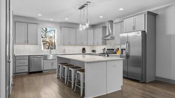 A modern kitchen with grey cabinets, a stainless steel refrigerator, a large island with bar stools, and a window providing a view of trees.