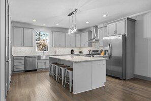 A modern kitchen with grey cabinets, a stainless steel refrigerator, a large island with bar stools, and a window providing a view of trees.