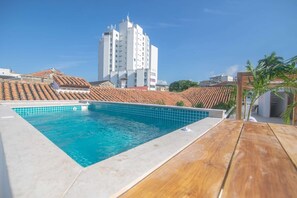 En swimmingpool p å taget med en træterrasse, omgivet af bygninger med røde tegltage.