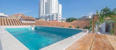 Una piscina en la azotea con un deck de madera, rodeada de edificios con techos de tejas rojas.