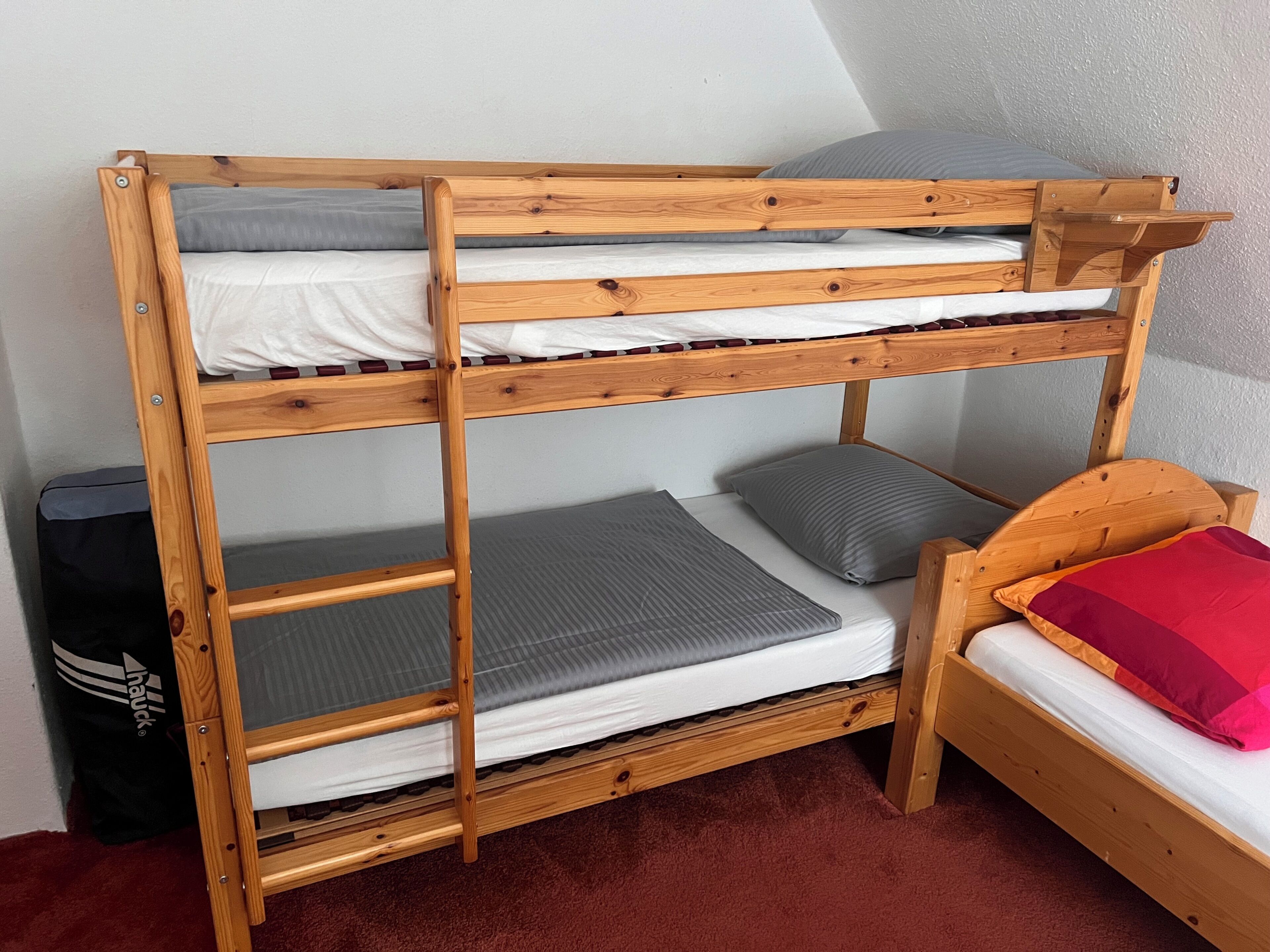 Ein Etagenbett mit Holzrahmen, grau-weißer Bettwäsche und einem roten Kopfkissen.