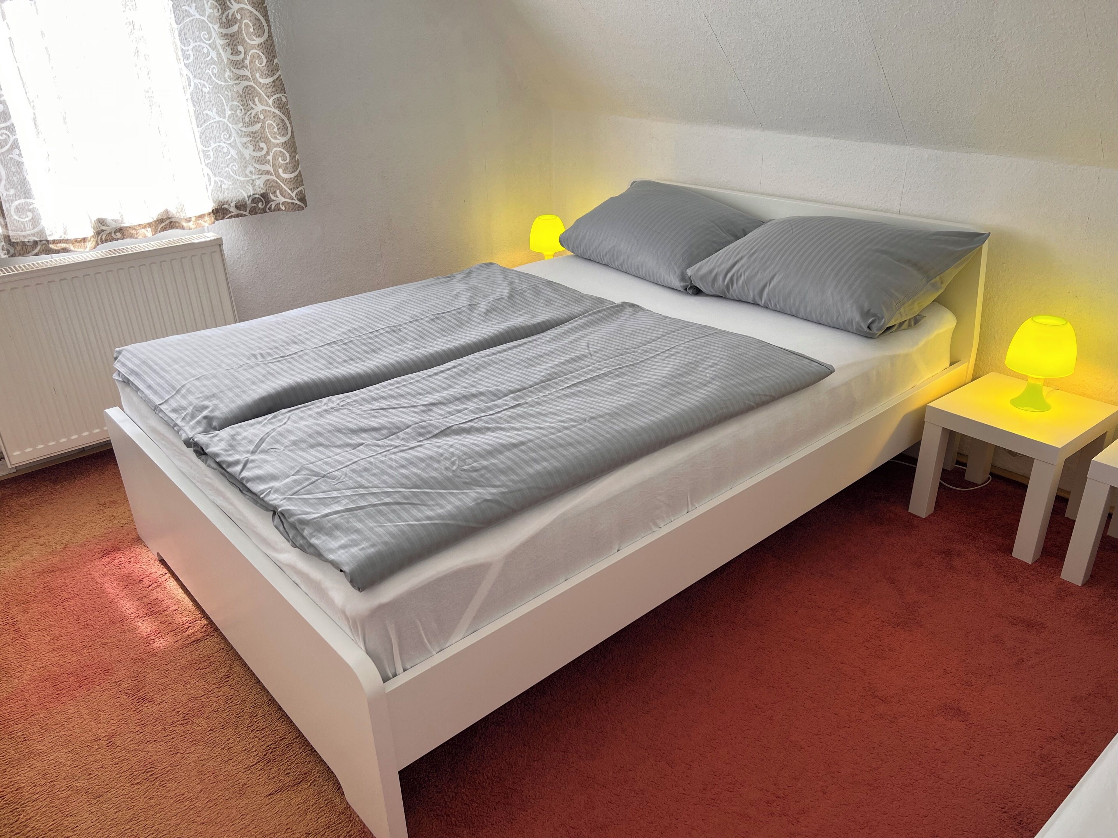 Una cama con dos almohadas, una mesita de noche con una lámpara y una ventana con cortinas.