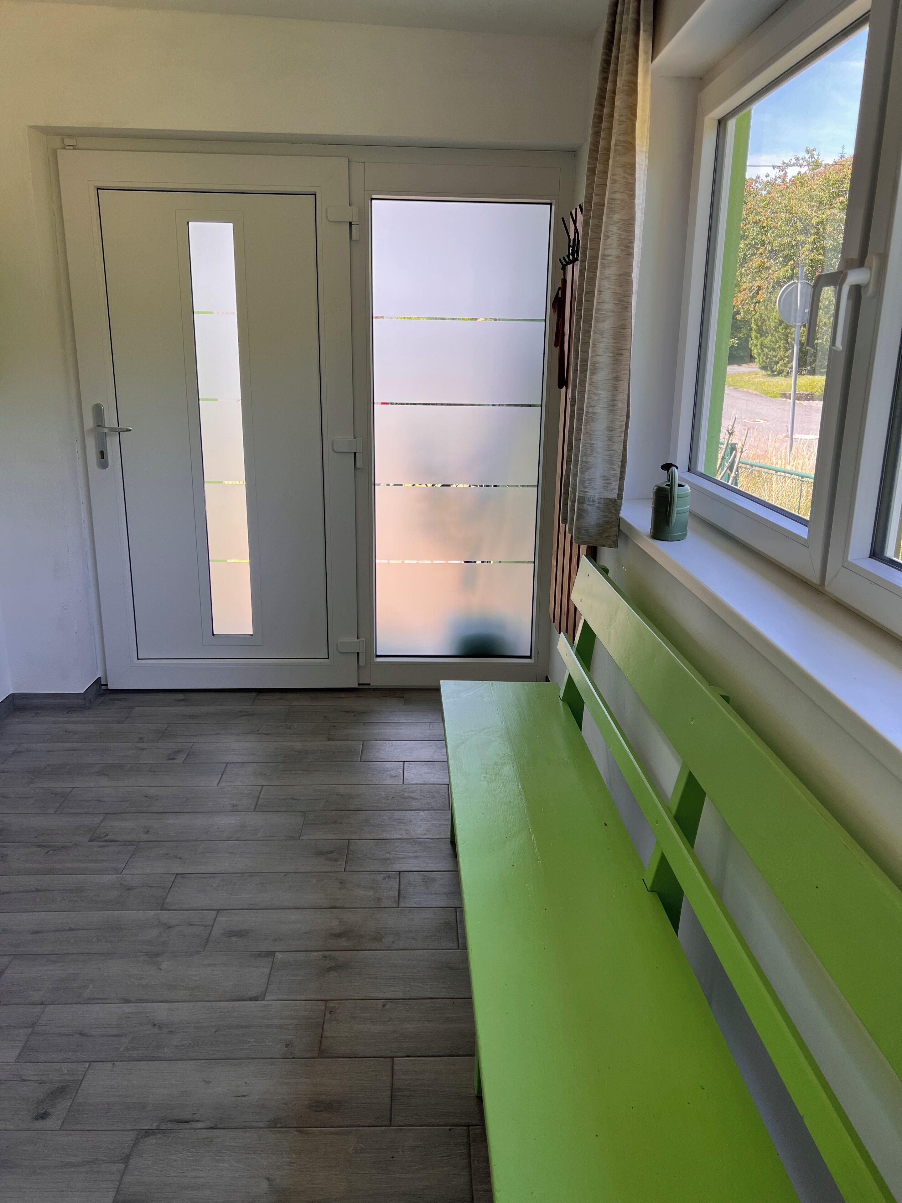 Ein Zimmer mit Holzboden, einer grünen Bank, einer weißen Tür mit satinierten Glasscheiben und einem Fenster mit Blick auf Grünflächen.
