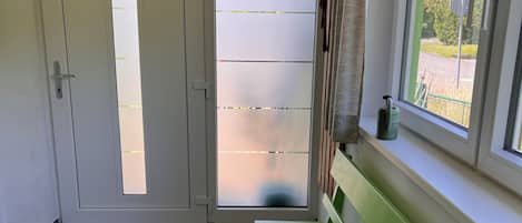 Una habitación con suelo de madera, un banco verde, una puerta blanca con paneles de vidrio esmerilado y una ventana con vistas a zonas verdes.