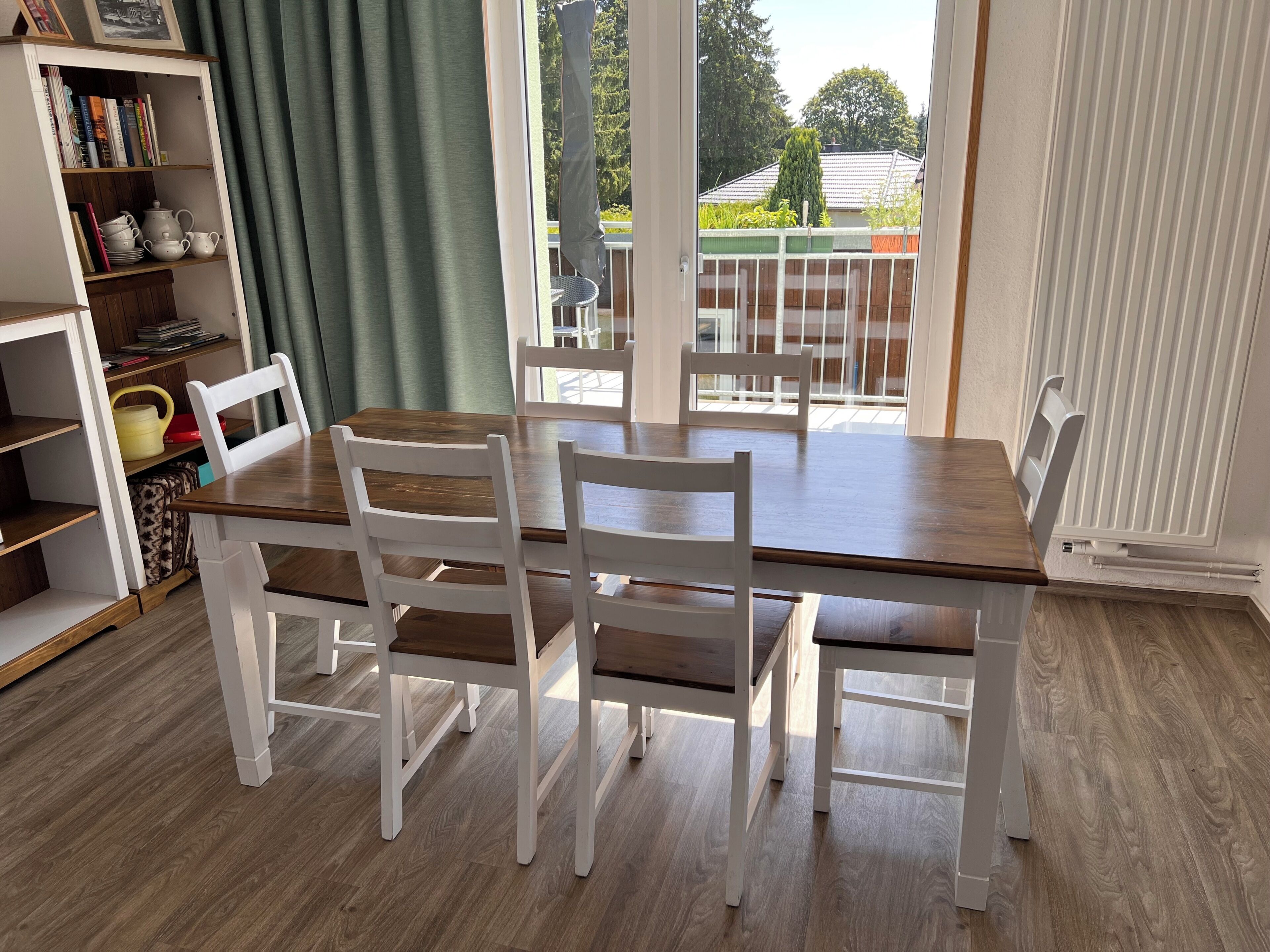 Ein Essbereich mit einem Holztisch und weißen Stühlen, ein Bücherregal mit Büchern und Dekoration, sowie ein Blick ins Freie durch große Fenster.