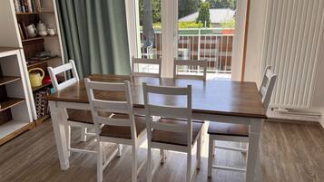 Ein Essbereich mit einem Holztisch und weißen Stühlen, ein Bücherregal mit Büchern und Dekoration, sowie ein Blick ins Freie durch große Fenster.