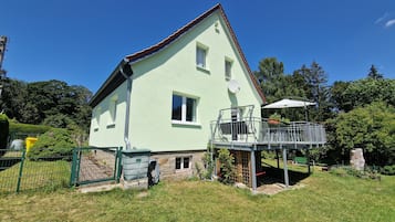 Una casa verde clara con balcón, rodeada por un patio cercado y árboles.