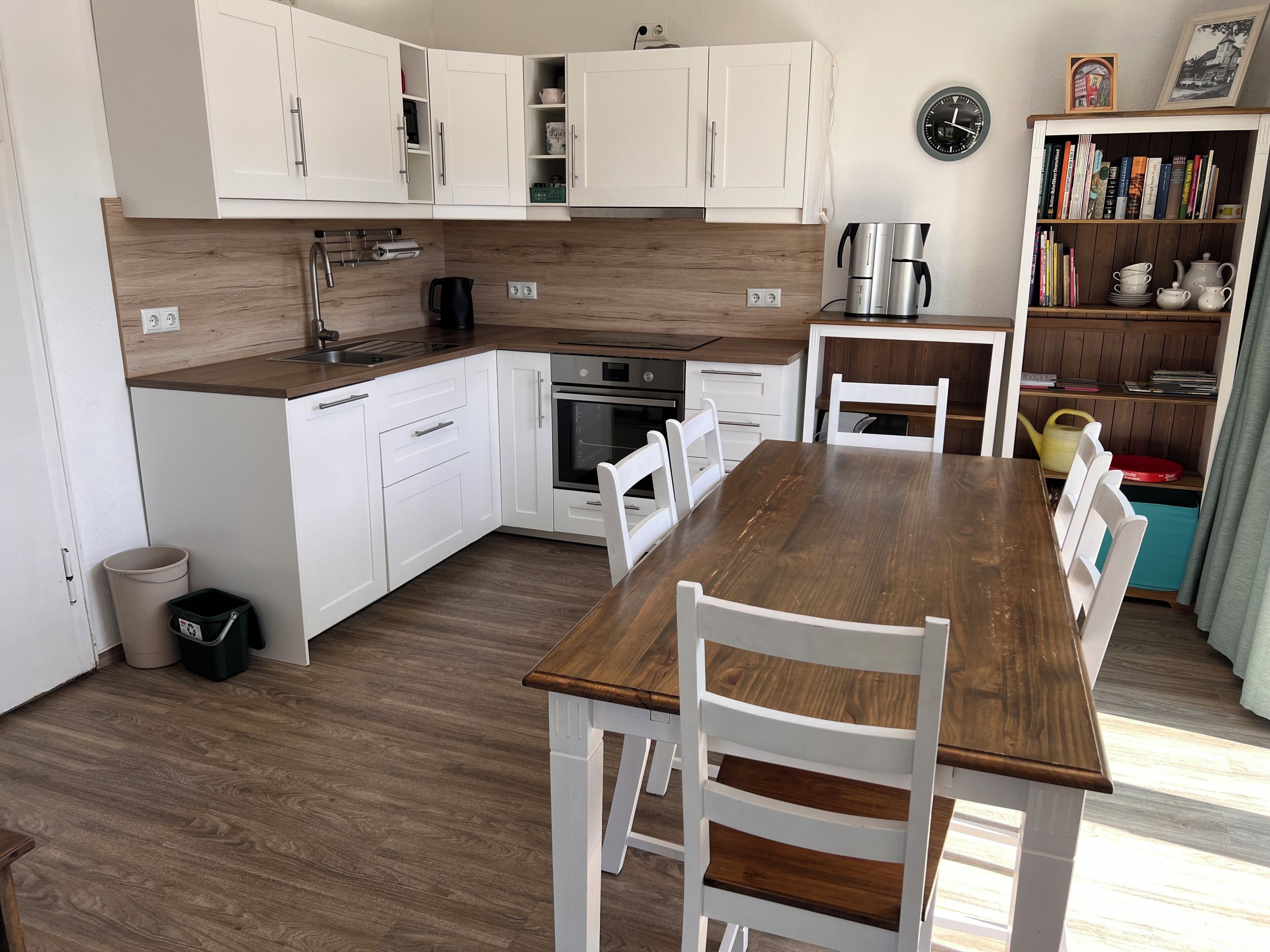 A modern kitchen with white cabinetry, a wooden countertop, a dining table with chairs, and a bookshelf with books and kitchenware.