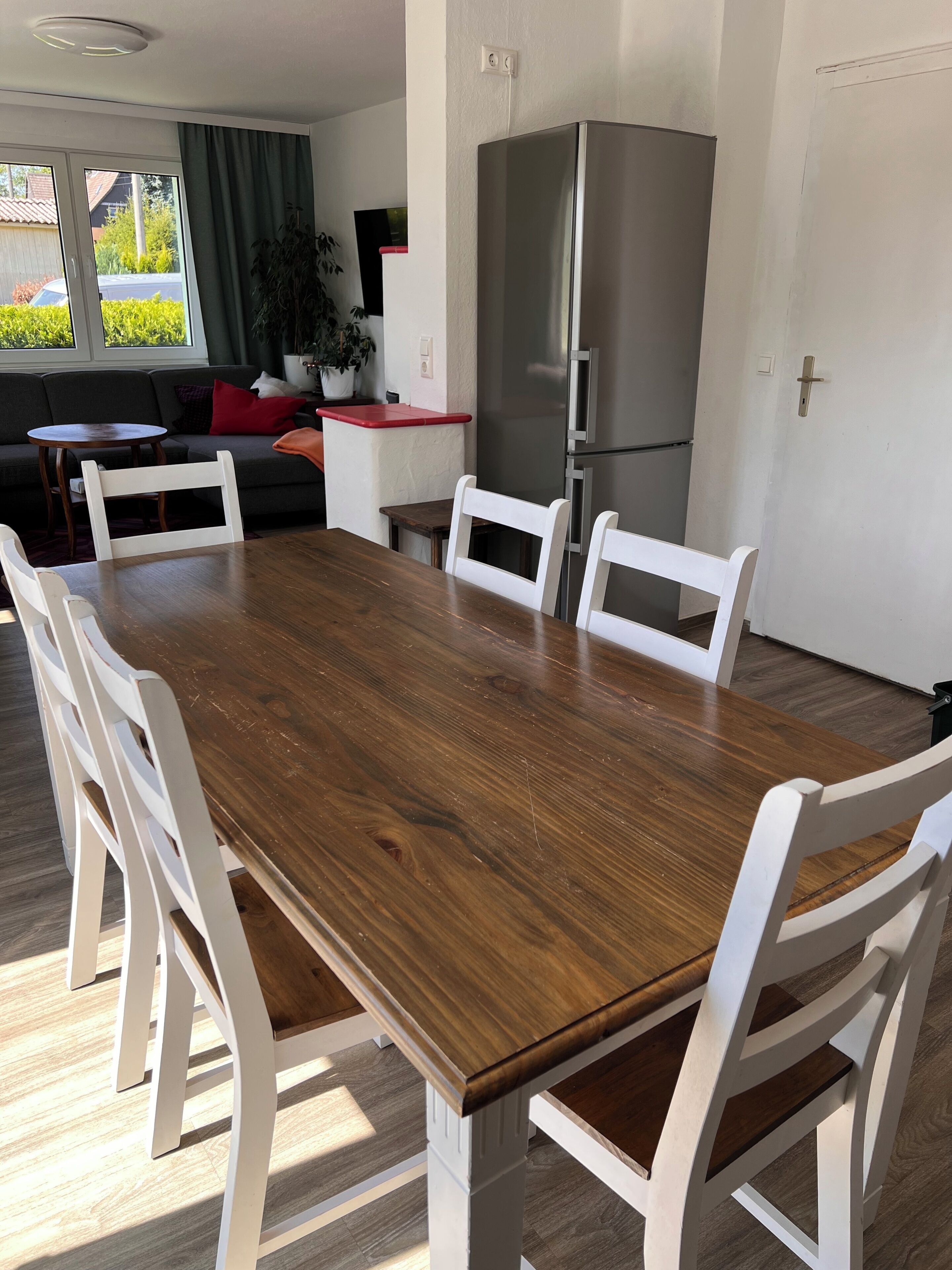 A dining area with a wooden table and white chairs, a refrigerator, and a sofa in the background.