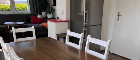 A dining area with a wooden table and white chairs, a refrigerator, and a sofa in the background.