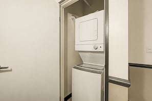 A compact laundry room with a washer and dryer, a door, and a wall-mounted light fixture. - New Stylish Indy Apt- Perfect for Groups - 3BR (Indianapolis)