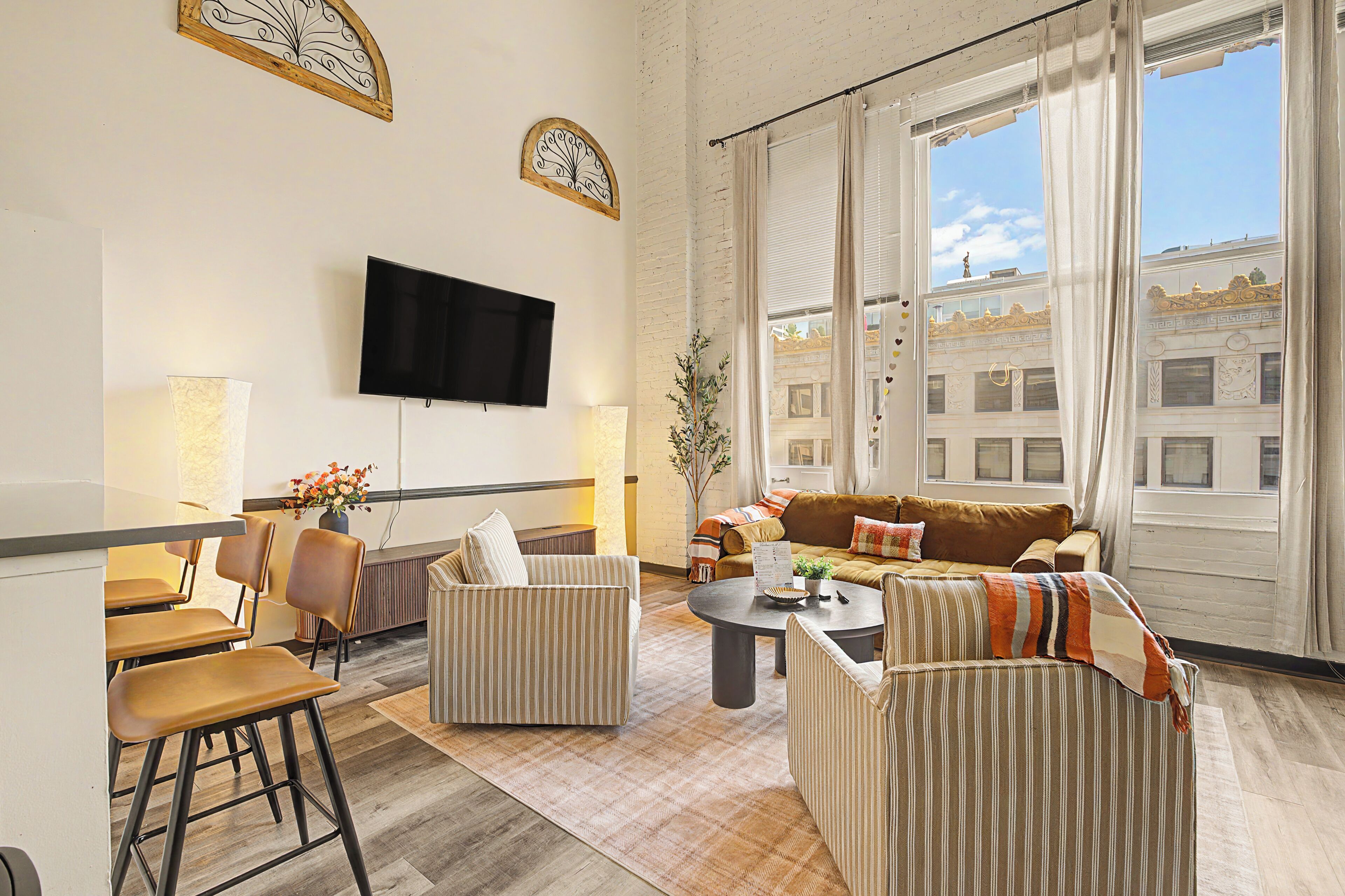 A modern living room with a flat-screen TV, a sofa set, a dining area with chairs, and a large window with curtains.
