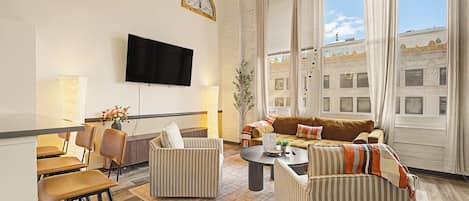 A modern living room with a flat-screen TV, a sofa set, a dining area with chairs, and a large window with curtains.