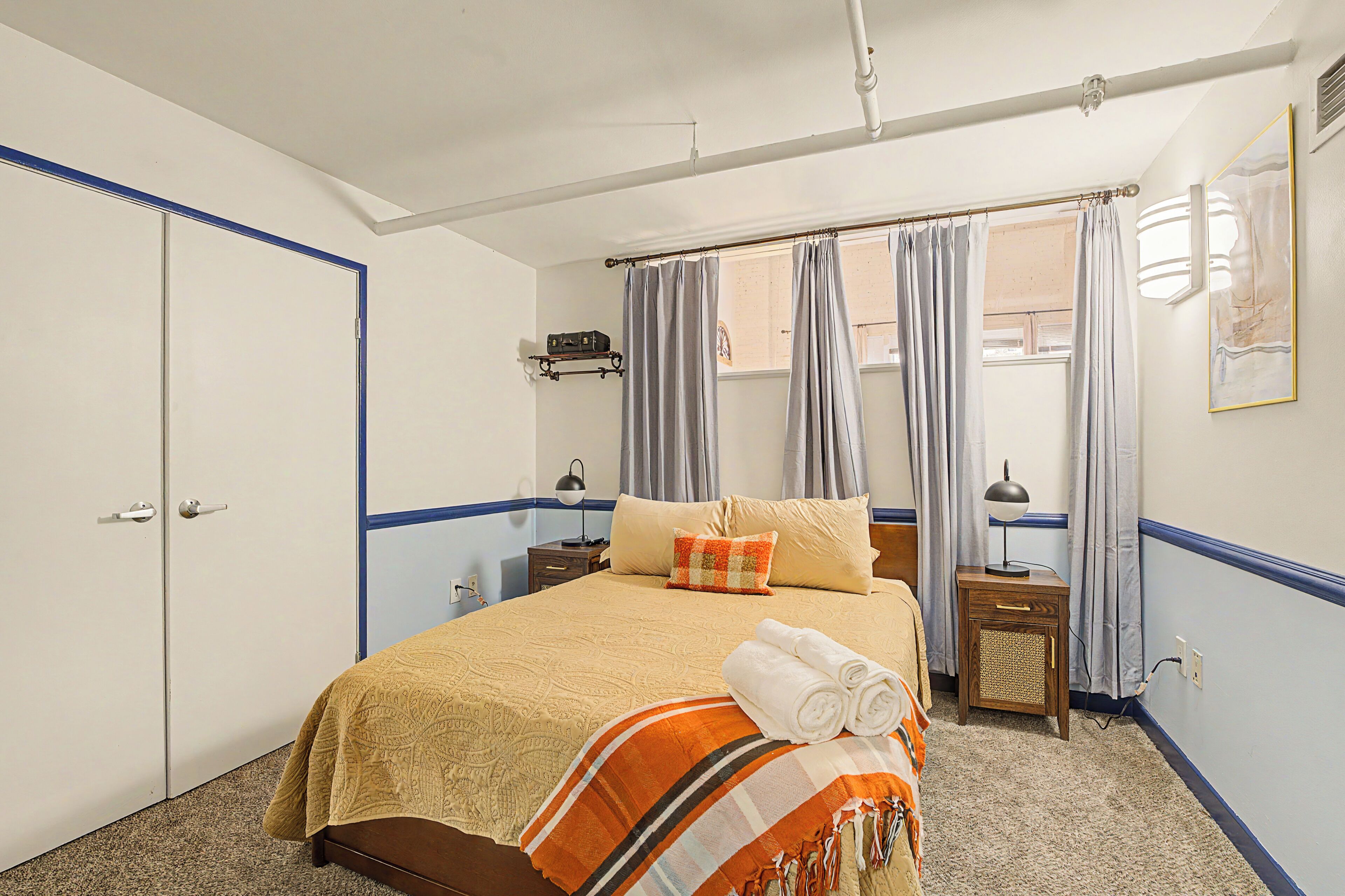 A hotel room with a bed, a nightstand, a lamp, a window with curtains, and a door.