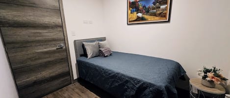 A bedroom with a bed, a wooden wardrobe, a framed picture on the wall, and a small round table with a plant.