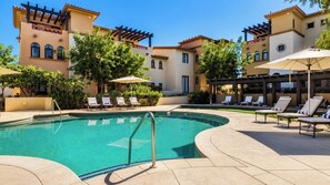 Ein Swimmingpool mit Liegestühlen und Sonnenschirmen, umgeben von Gebäuden im mediterranen Baustil.