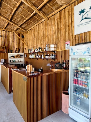 Zona de bar con pared de bambú, mostrador de madera y un refrigerador lleno de bebidas.