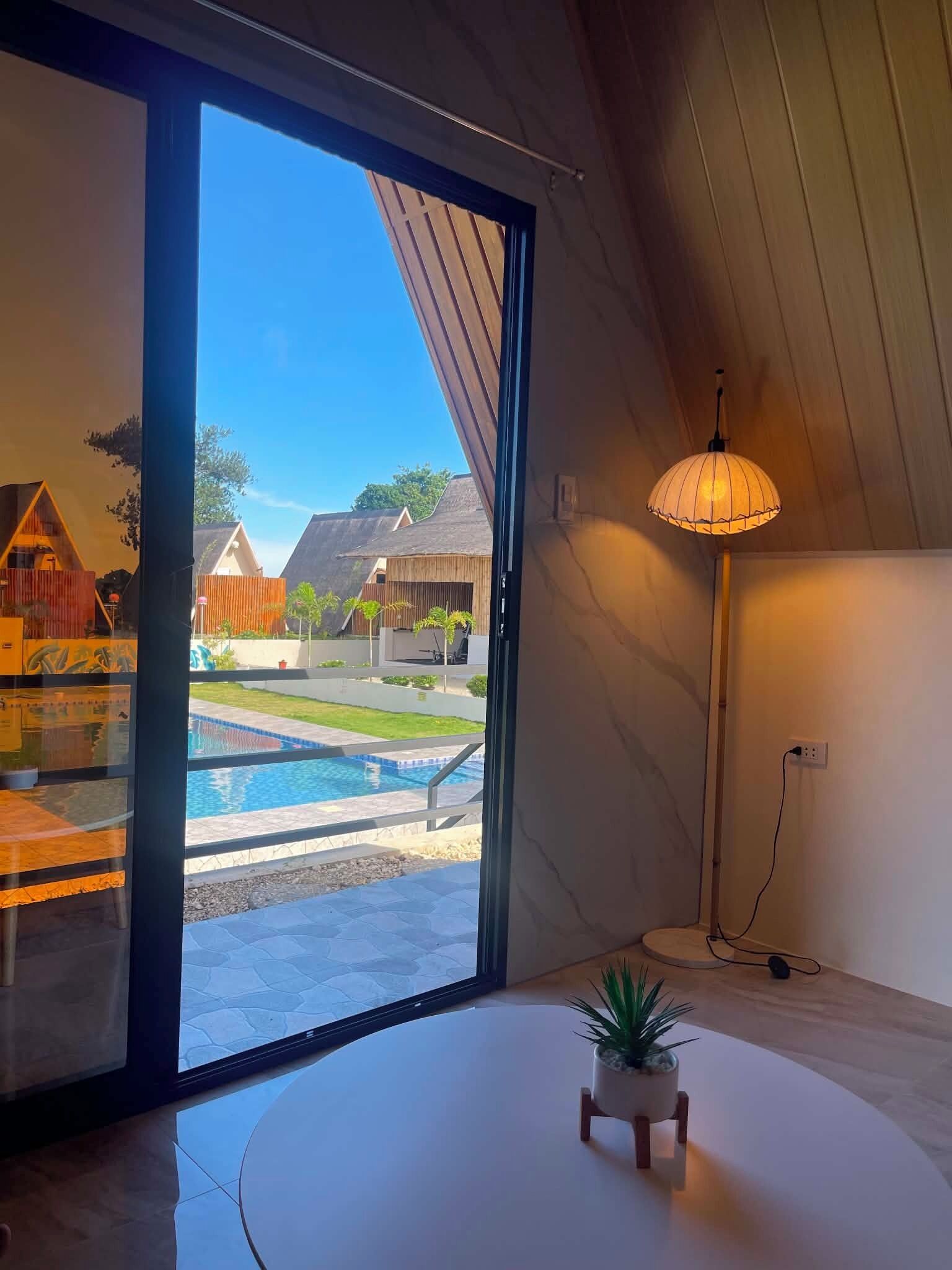 A room with a sliding glass door leading to a pool area, a small potted plant on a white table, and a floor lamp.