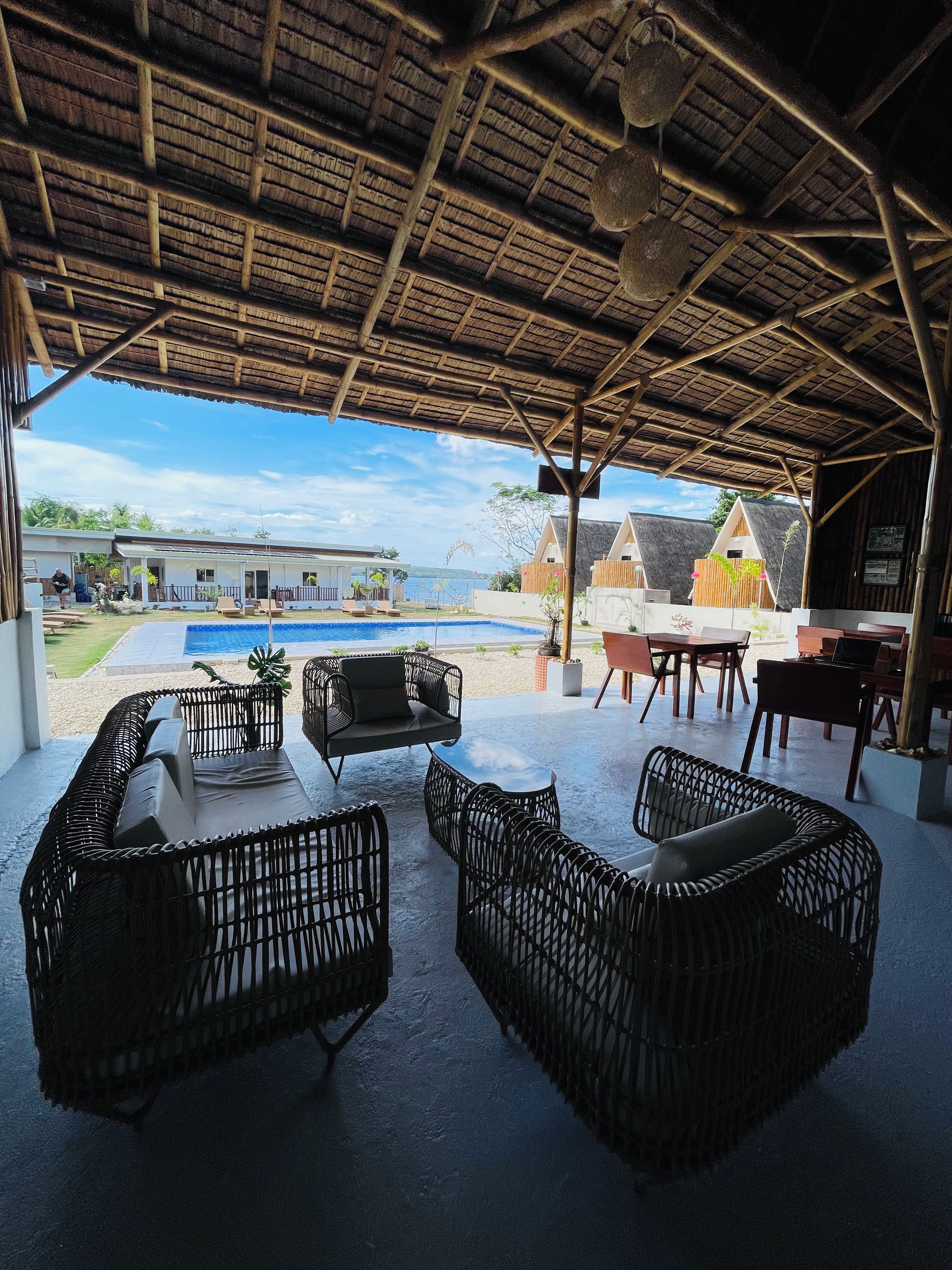 A covered outdoor seating area with wicker furniture, a pool, and a view of a resort with thatched-roof buildings.