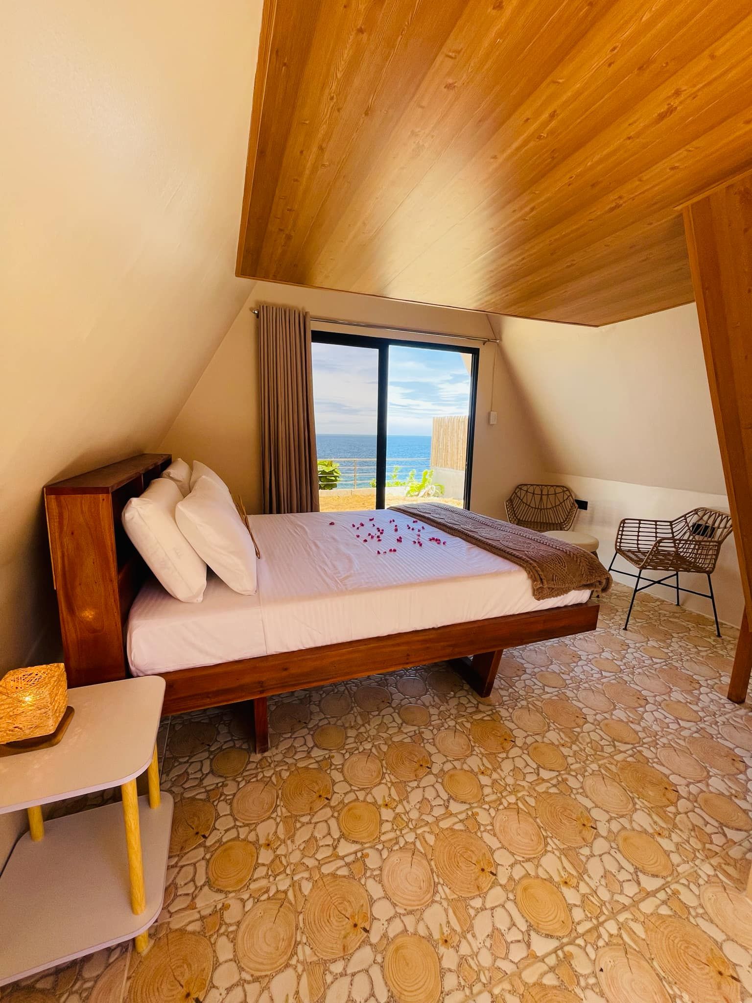 A bedroom with a wooden ceiling, a bed with white bedding, a view of the sea, and a stone-patterned floor.