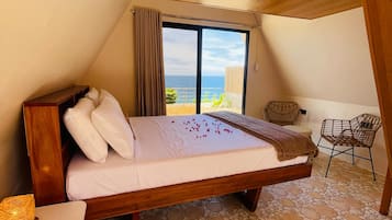 A bedroom with a wooden ceiling, a bed with white bedding, a view of the sea, and a stone-patterned floor.