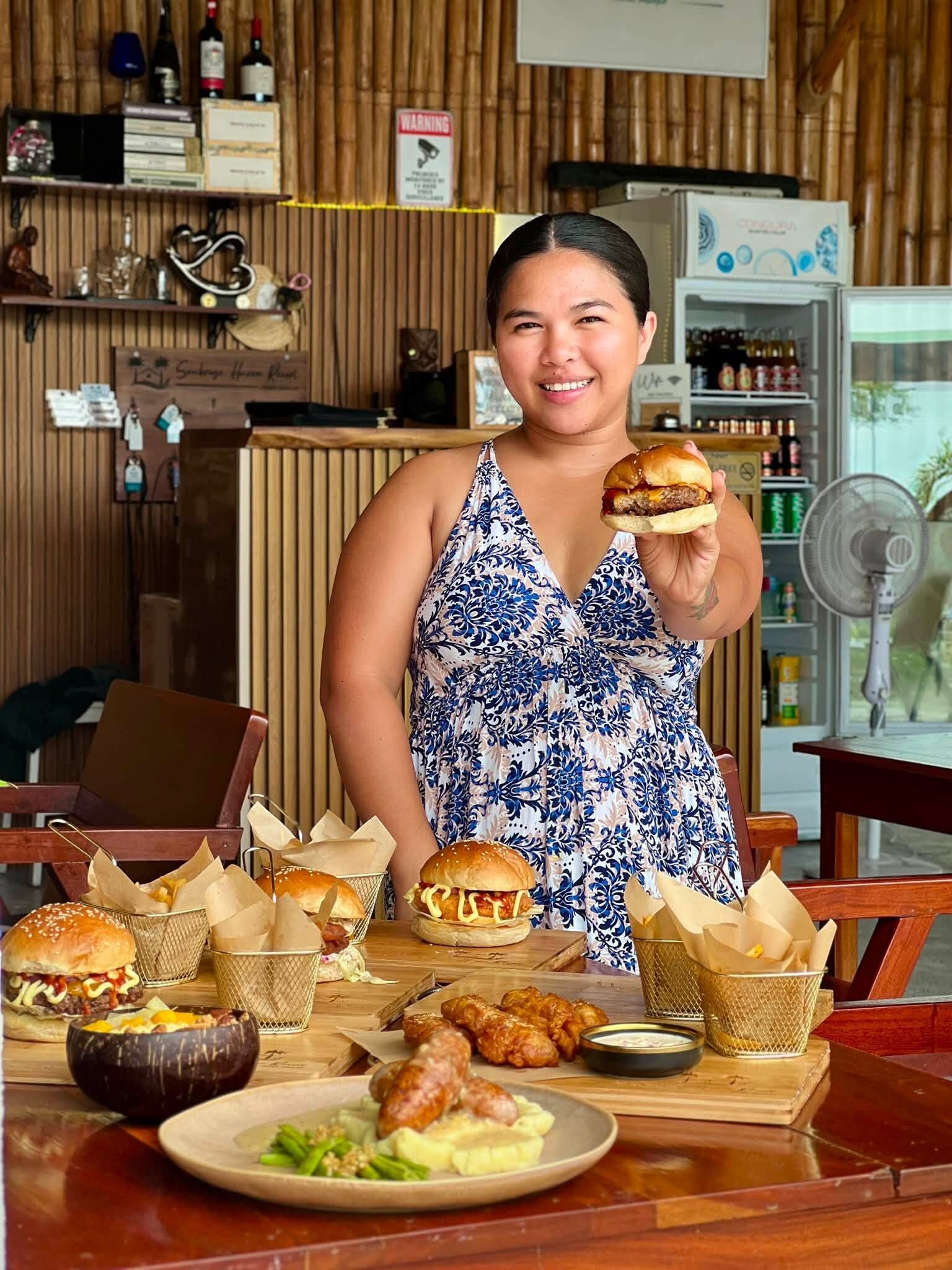 A person holding a burger in a restaurant with a wooden interior, a refrigerator, and various food items on the table.
