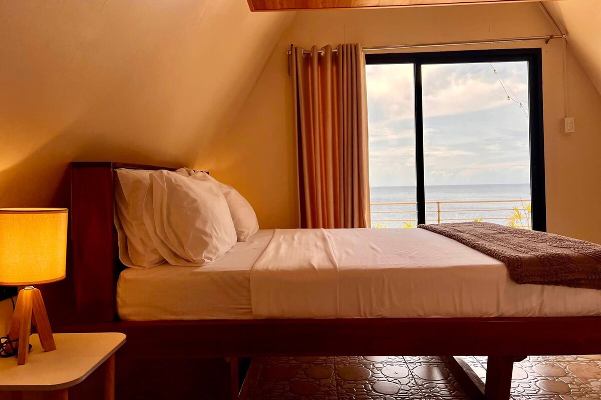 A bedroom with a bed, a nightstand, a lamp, a window with curtains, and a view of the sea.