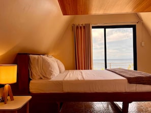 A bedroom with a bed, a nightstand, a lamp, a window with curtains, and a view of the sea. - Seabreeze Haven Resort (Maria)