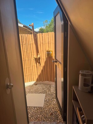 Un baño con puerta de madera, zona de ducha con suelo de baldosas y un pequeño estante con un recipiente blanco.
