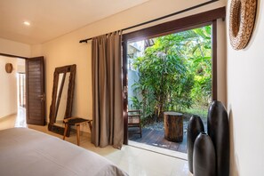 A bedroom with a large bed, a wooden chair, a small table, and a view of a garden through a sliding door. - Private Pool | Garden Lounge | Dedicated Workspace (Canggu)