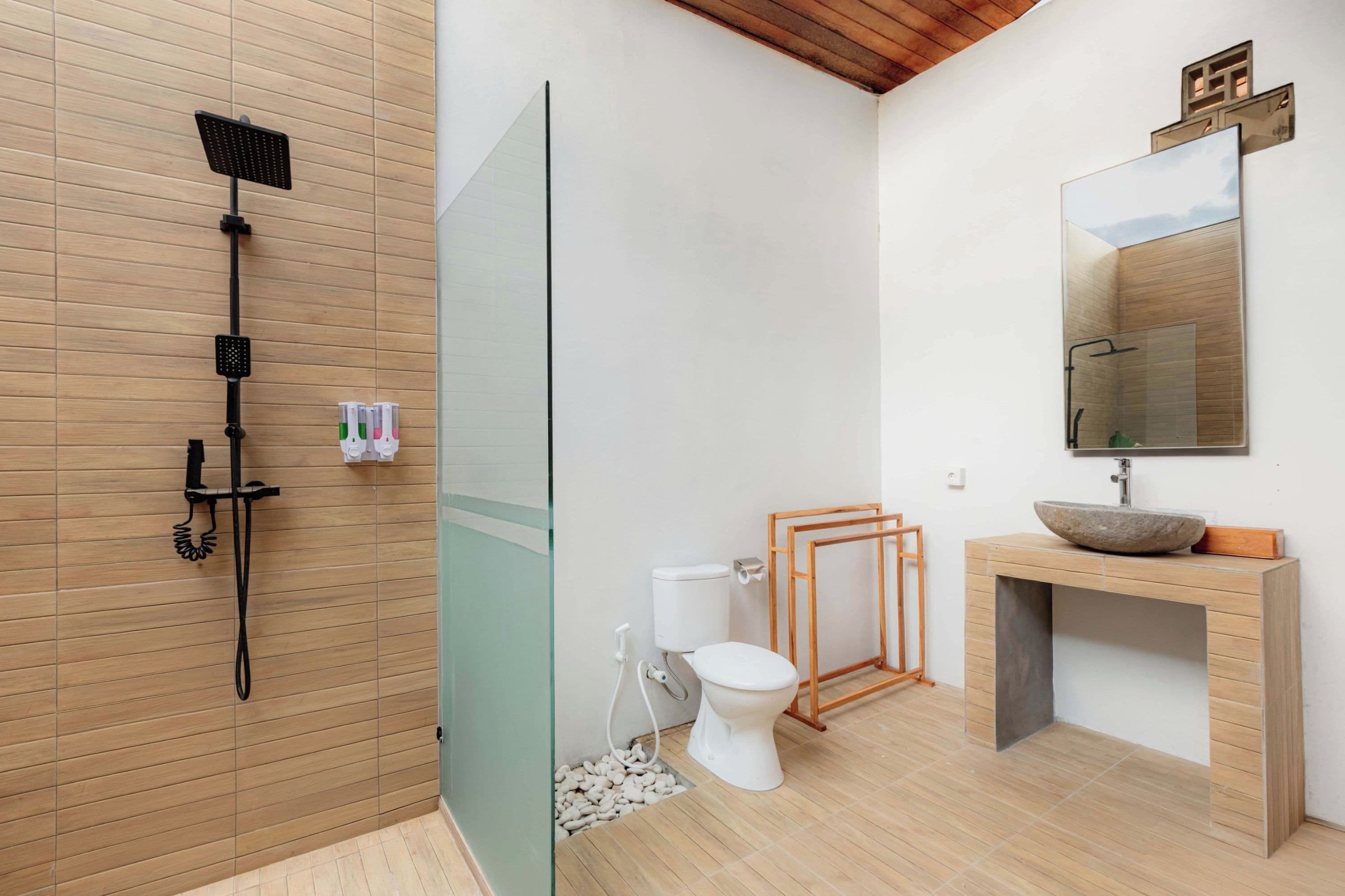 Un baño moderno con piso de madera, cabina de ducha de vidrio, inodoro blanco y tocador de madera con lavabo de piedra.