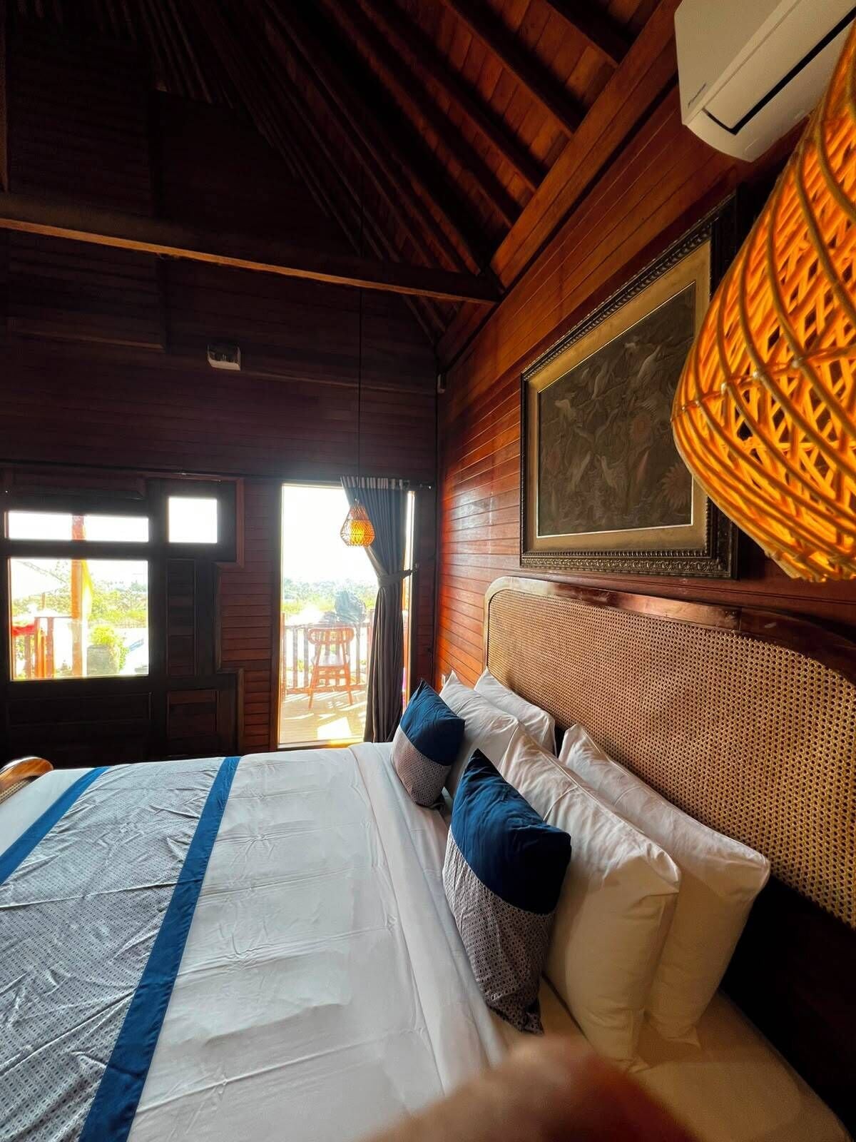 A hotel room with a bed, a large window, a ceiling fan, and a framed picture on the wall.