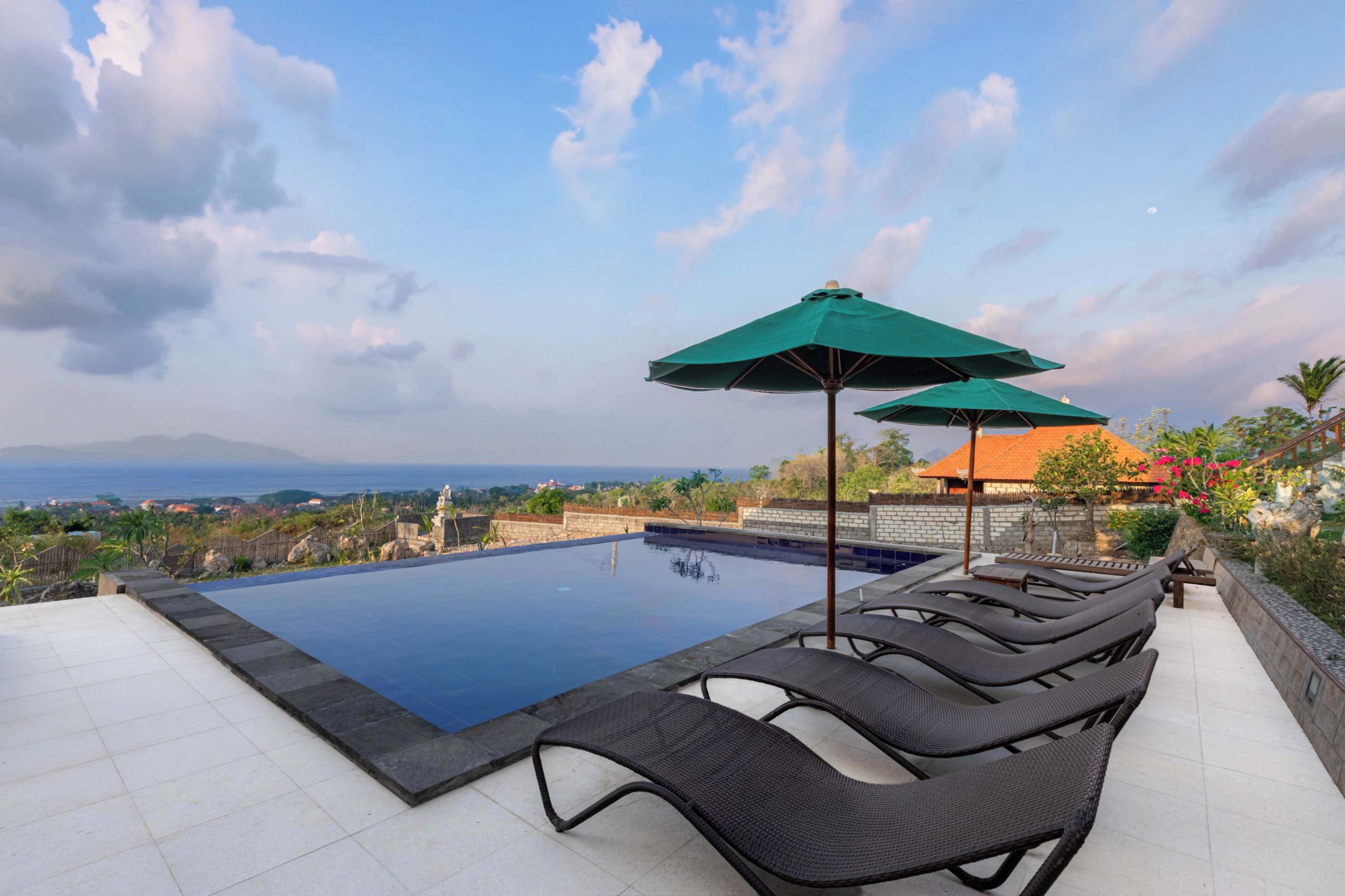 Una piscina con sillas de descanso y sombrillas, rodeada de un jardín ajardinado y vista al océano.