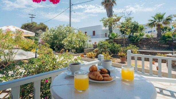 A table with breakfast items including croissants, coffee, and orange juice, set against a backdrop of a garden with various plants and a white building. - NOSTOS SIFNOS (Apollonia)
