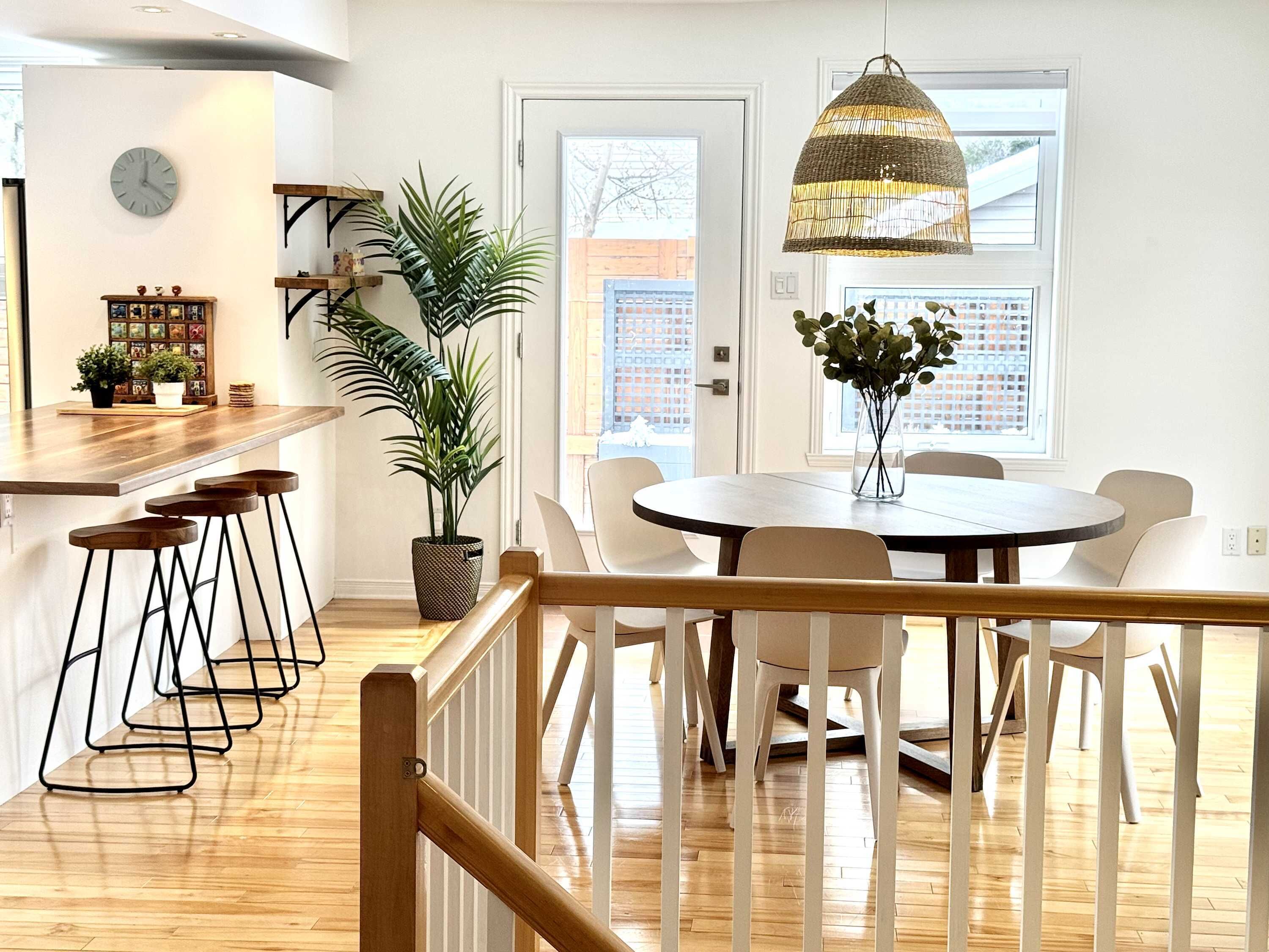 A modern kitchen with a wooden countertop, bar stools, a dining table with chairs, a hanging pendant light, and a potted plant.