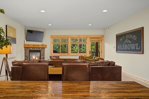 Ein gemütliches Wohnzimmer mit Kamin, einem großen Holzesstisch, bequemen Ledersofas und Blick ins Freie durch große Fenster.