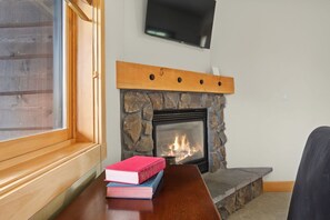 A fireplace with a stone surround, a television mounted above it, and a window with blinds to the left. - Wy’East Condo 104 at South Grand Lodge | Dog Friendly, Best Mt. Hood View (Government Camp)