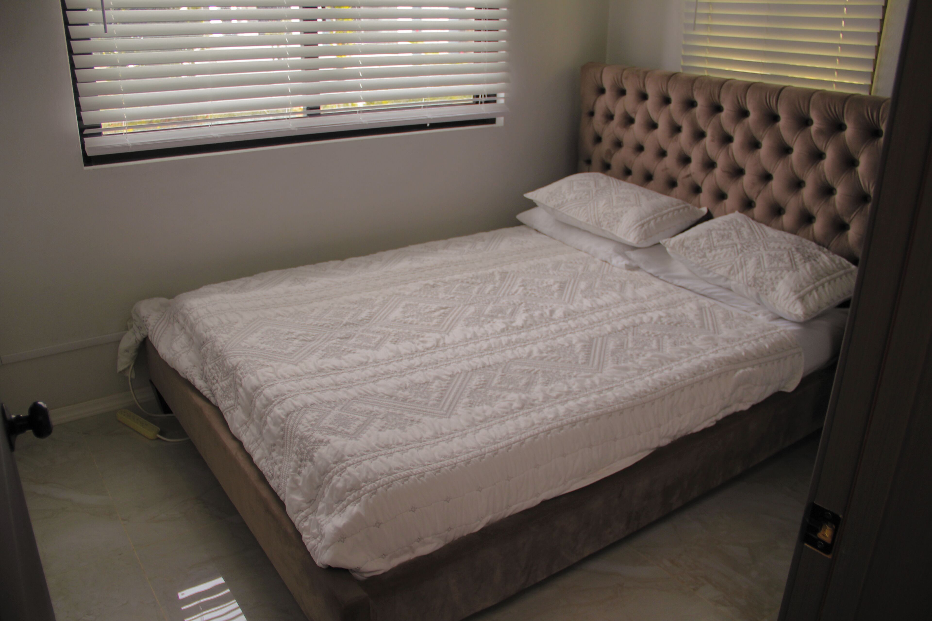 A bedroom with a single bed, a window with blinds, and a wall-mounted light switch.