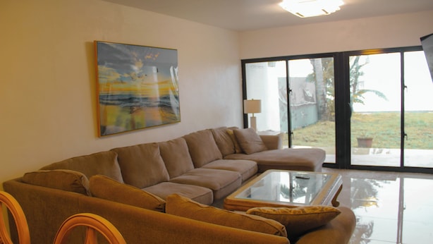 A living room with a sectional sofa, a glass coffee table, and a painting on the wall. - Beachfront home with beautiful sunset views. Must see! (Asan)