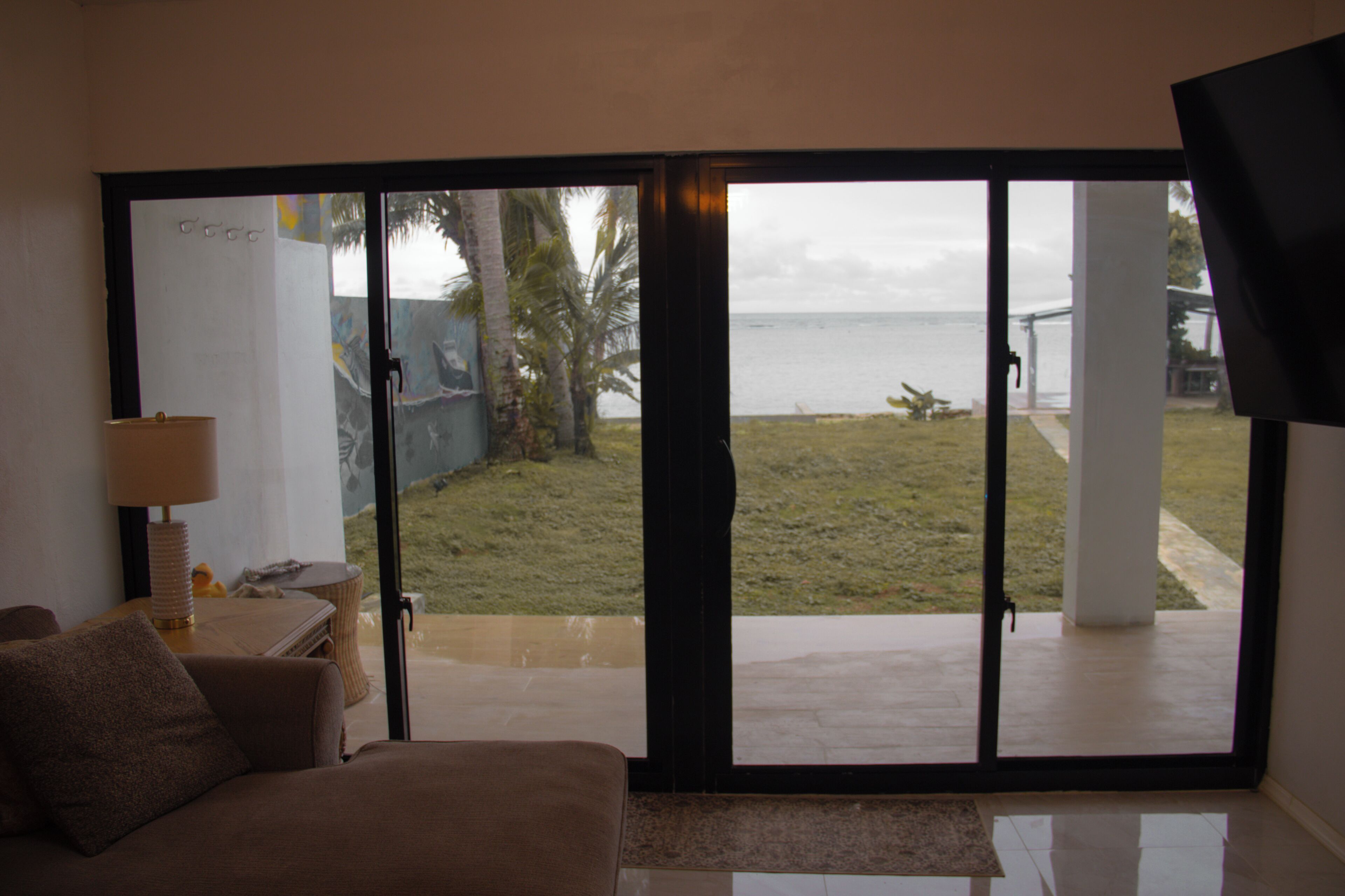 Una habitación con sofá, lámpara y vistas a través de un ventanal corredero: un espacio verde con palmera y un cuerpo de agua.