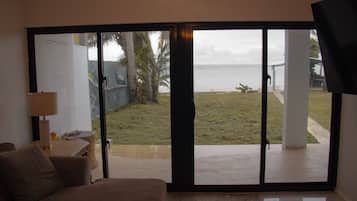 Una habitación con sofá, lámpara y vistas a través de un ventanal corredero: un espacio verde con palmera y un cuerpo de agua.