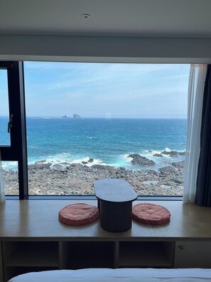 Habitación con una ventana grande con vistas a un litoral rocoso y al mar. Cuenta con una cama con ropa blanca, una mesita con dos cojines rosados y un taburete oscuro.