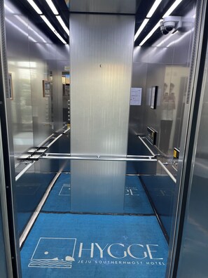 Interior de ascensor con paredes metálicas reflectantes, alfombra azul con el logotipo de un hotel y una cámara de seguridad.
