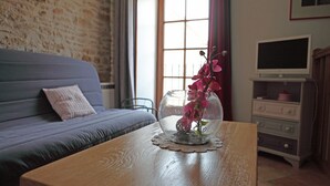 Ein Zimmer mit einer Couch, einem Holztisch mit einer Glasvase und Blumen, einem Fernseher auf einem Schrank und einem Fenster mit Vorhängen.